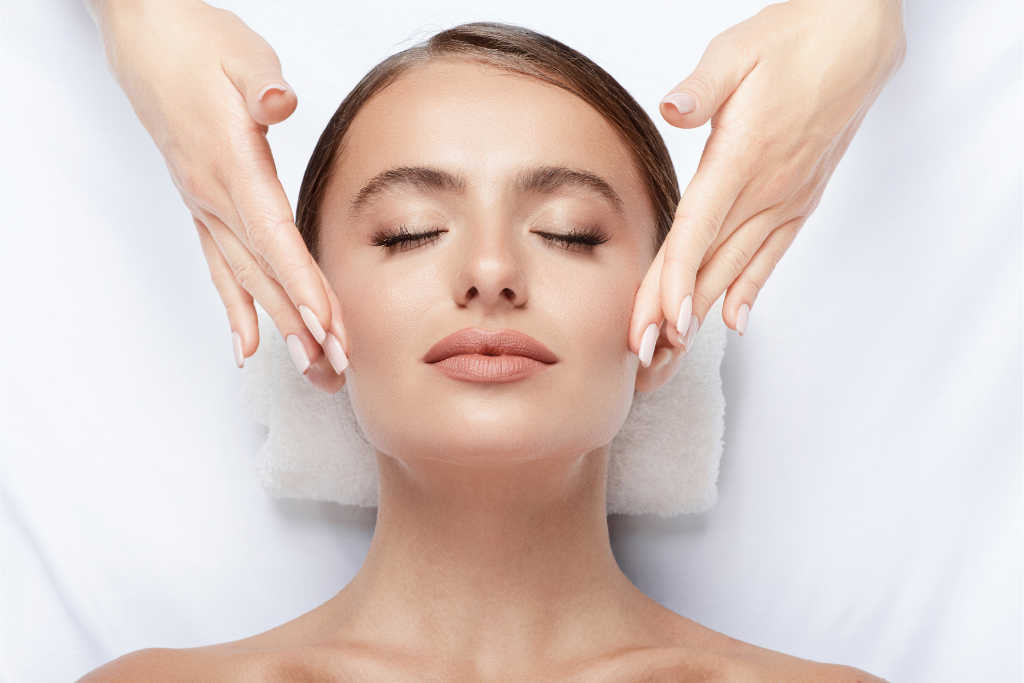 A woman with closed eyes lying on a white towel receives a relaxing facial massage from two skilled hands.