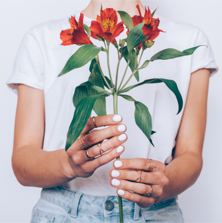 Person wearing white nail polish and rings, holding a bouquet of red flowers with green leaves. They are dressed in a white t-shirt and blue jeans—just one of the stylish moments from Peachy Peel beauty salon in London, where you can find various packages and offers.